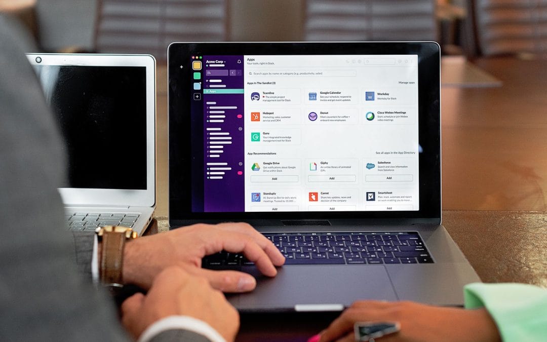 Two people sit at a table with laptops. One laptop displays Slack’s app directory, highlighting integrations popular among South African HR software users. One person’s hand is on the trackpad and the other’s hand holds a pen. In the background are empty office chairs and a conference table.